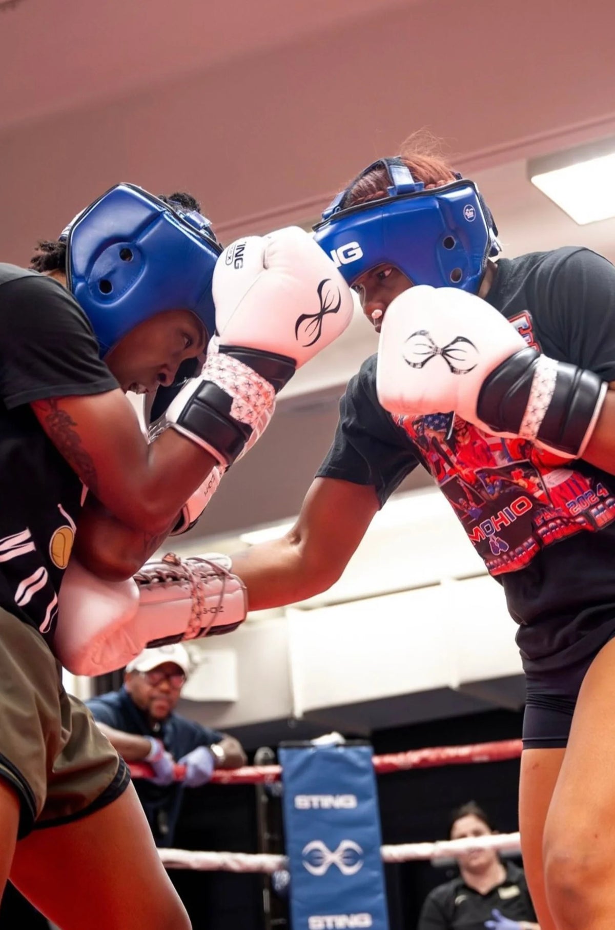 Two boxers in a boxing ring with one wearing white sting gloves and the other in black, with a referee in the background.