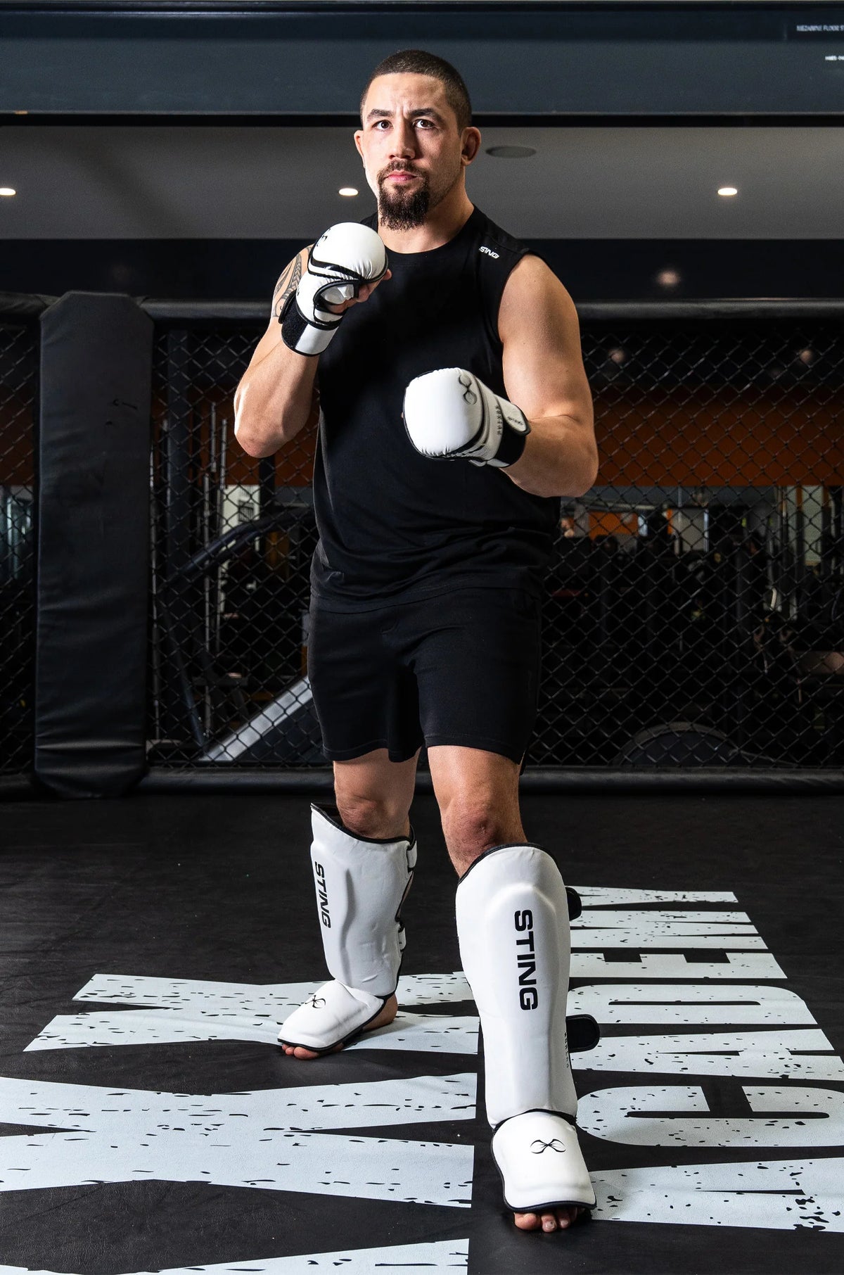 Man in black athletic wear with white boxing gloves and shin guards standing in a fighting stance on a branded mat.