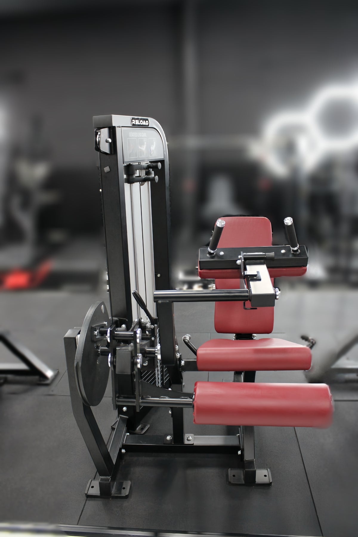 Leg curl machine in commercial gym setting. Reload By Fitness Hero. Matte black frame with a red seat