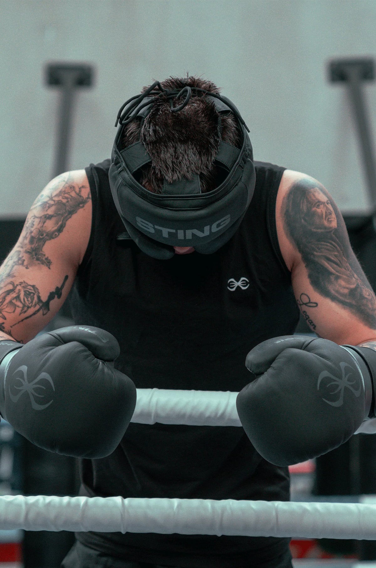 Person wearing a black tank top with 'STING' branding, gloves, and a mask in a boxing ring.